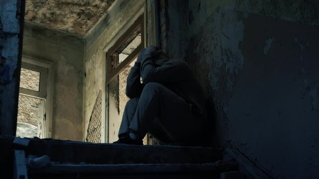 A young guy on the steps of an abandoned house in depression, a sad man, a person has social problems. The boy is wearing a hoodie in sadness. Earthquake destruction of the house.
