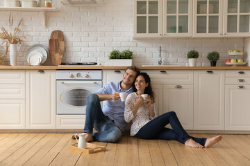 Homeowners family couple in love relax on wooden warm floor in kitchen holds cups drinking tea or...