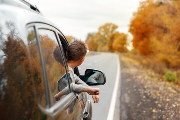 Boy putting his heads and hands out of the car window driving down a country autumn road . Motion blur. Kid enjoying view of the road from the car. Feeling freedom, travelling concept. 