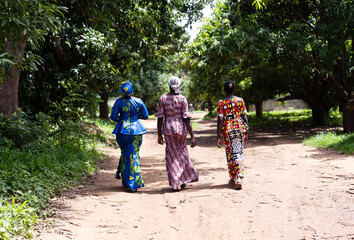 Obraz premium Rear view of three African women walking long distances on a dirt road due to a lack of transport links; missing infrastructure concept