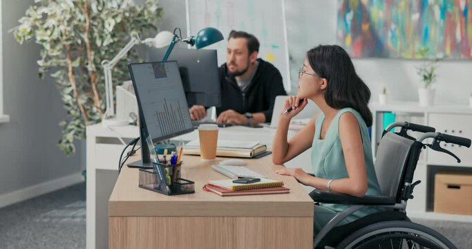 Workday in office, corporation, company, co-workers are working on computers, drafting documents, woman in glasses with Asian beauty is sitting at desk in wheelchair, she is disabled, after accident