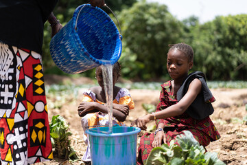 Black African girls sitting in a dry field, with a bucket about to be filled with water; water...