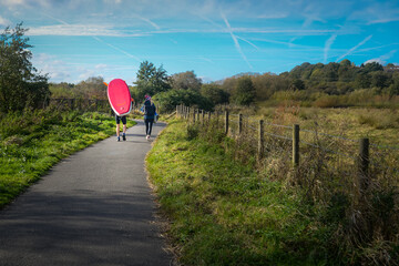 Two people with paddle board walk along country path towards river.
