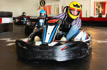 Glad cheerful smiling male in helmet and other people driving cars for karting in sport club indoor - obrazy, fototapety, plakaty