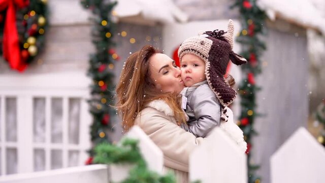 Beautiful mother kisses a little girl in a bull's hat against the background of a Christmas house under the flying snow. Solw motion
