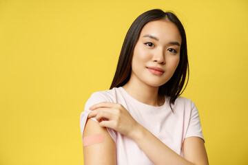 Covid-19 and healthcare medical concept. Portrait of asian female shows her shoulder with patch after coronavirus vaccination, yellow background - obrazy, fototapety, plakaty