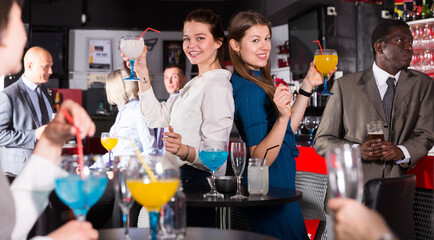 Cheerful girls with coworkers funning in bar during firms party, dancing and toasting drinks