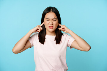 Annoyed asian girl shut ears and grimacing, loud sound, displeased by siren, standing in t-shirt...