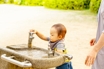 公園の水飲み場（手洗い場）で水遊びする子供。後ろでママが見守っている。または手を洗おうとしている子供