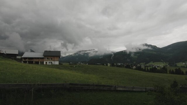 Timelapse of storm clouds rolling into a valley with mountains in the background. A wooden challet and green grass in the foreground.