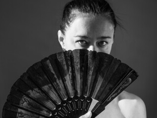 Portrait of beautiful attractive full face brunette woman with fan covering her face, bare shoulders. Black and white photo on black background.