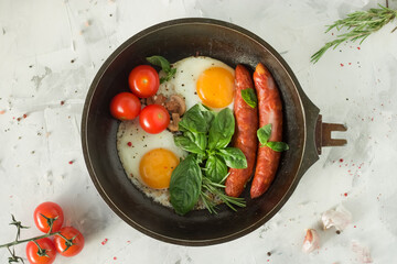 Frying pan with two fried eggs, cherry tomatoes, basil and sausages on a light background.