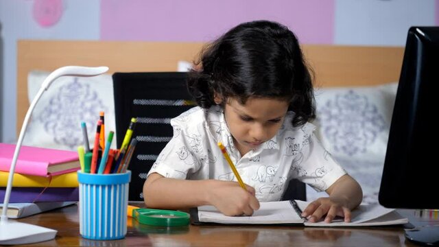 A small adorable child with curly black hair studying at home on a desk. Closeup shot of a young boy completing his homework in his room - distant education