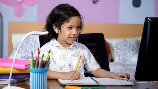 A Little Boy With Curly Black Hair Taking Online Classes During The  COVID - 19 Pandemic. Closeup Shot Of An Adorable Kid Wearing White Earphones Talking To His Teacher On A Video Call