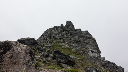 Fototapeta na wymiar The top of the cliff with bizarre outlines against the foggy sky. Cracks on the rocks and sparse vegetation. A tiny silhouette of a man on a peak is visible. Kamchatka. Mount Camel