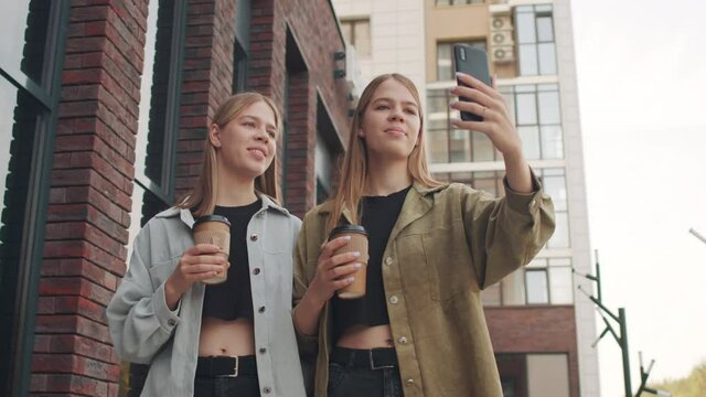 Low-angle tracking shot of young twin sisters with coffee in to-go cups laughing and recording video of themselves on mobile phone while walking down street together