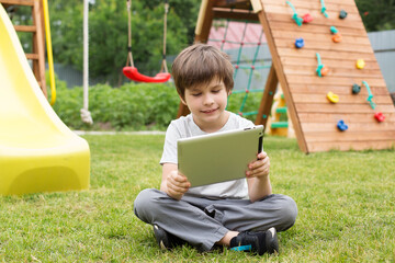 a a smiling boy with a tablet is sitting on the green grass on the playground. Online training, online games