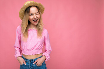 Photo shot of young beautiful cute happy blonde woman wearing stylish pink crop top and straw hat isolated over pink background with copy space