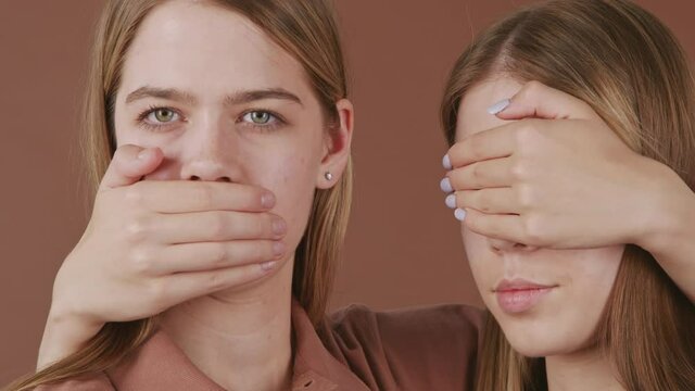 Panning close up portrait shot of young twin women covering mouth and eyes while posing for camera against brown background