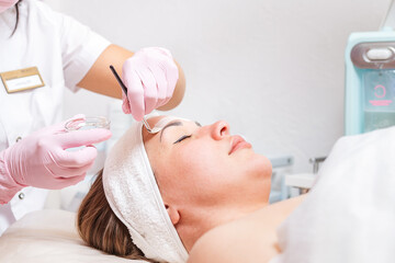 Obraz na płótnie Canvas Beauty salon. The cosmetologist in medical gloves holds a bowl with a mask, applying cosmetics with a brush on the client's face. Copy space. Close up.Concept of professional cosmetology and treatment