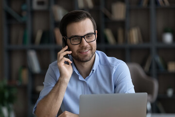 Happy friendly young business man manager employee in eyewear holding phone call conversation, giving professional consultation to client working on computer in modern home office, multitasking.