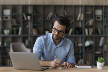 Concentrated young man in eyeglasses wearing wireless headphones with microphone, listening educational online lecture or holding business negotiations video call meeting, writing notes in copybook.