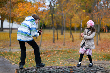 Obraz premium Children playing in autumn park. Girl and boy standing on the trunk of a tree and playing with a twig.