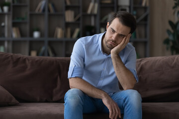 Bored millennial generation man sitting on sofa, feeling lonely at home. Unhappy young male suffering from negative thoughts, having depressive mood or problems in relations, regretting mistakes.