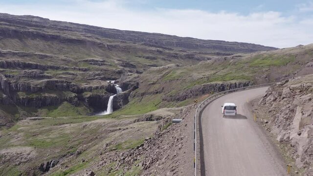 White car travels on dirt road passing scenic waterfall in Iceland, aerial
