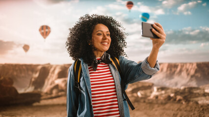 Excited Young Multiethnic Traveller with Afro Hairstyle Making a Selfie on a Smartphone and Showing...
