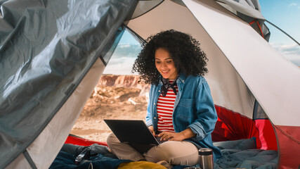Excited Beautiful Young Multiethnic Female Traveller with Afro Hairstyle Using Laptop Computer in a Tent on Top of Rocky Canyon. Adventurous Mountain Hiker Living in Nature in Great Outdoors.