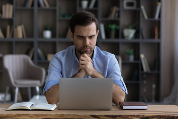 Pensive young businessman employee manager looking at laptop screen, considering difficult problem solution, thinking on online project, enjoying distant freelance workday in modern home office.