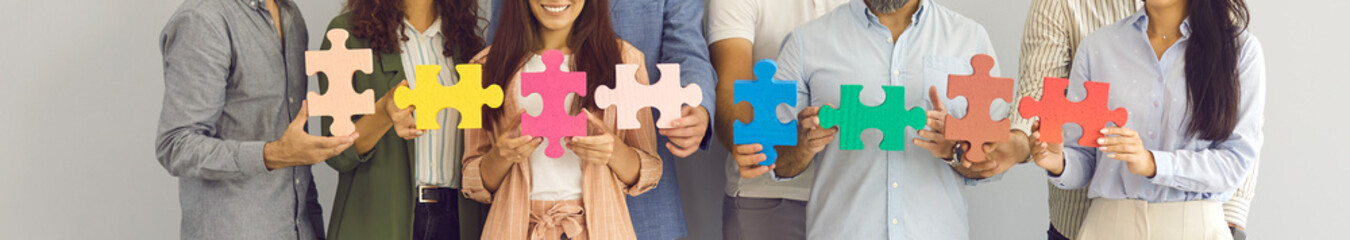 Team of happy business people making jigsaw chain. Banner background with group of entrepreneurs, coworkers and colleagues standing together and holding colorful jigsaw puzzle pieces. Teamwork concept - obrazy, fototapety, plakaty
