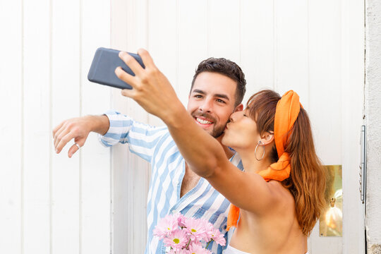 Girlfriend taking selfie through smart phone while kissing boyfriend by white wall