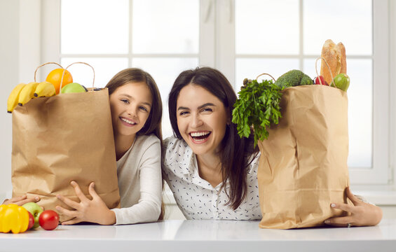 Happy Family Unpacking Groceries At Home. Portrait Of Cheerful Beautiful Mom And Kid At Kitchen Table Counter With Bags Full Of Apples, Oranges, Peppers, Tomatoes, Zucchinis, Parsley From Food Market