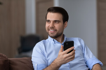 Obraz na płótnie Canvas Happy relaxed young handsome man looking in distance holding cellphone in hands, thinking or received message with pleasant news, dreaming resting on comfortable sofa, spending free time online.