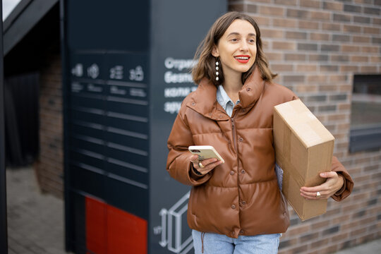 Young caucasian woman with parcel near automatic post terminal. Smiling girl holding smartphone and looking away on city street. Concept of smart delivery. Idea of modern shipping and logistics