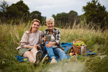 Naklejka na ściany i meble Senior mother and adult daughter sitting and having picnic outdoors in nature, looking at camera.