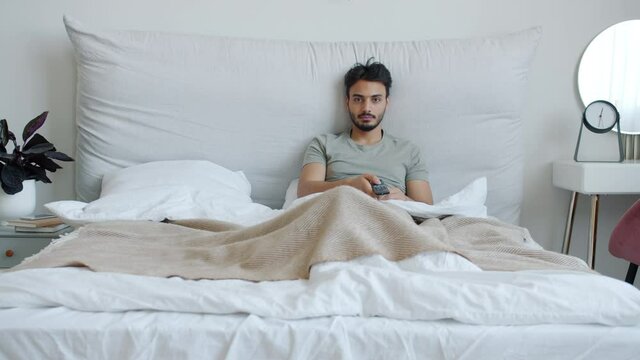 Happy Arab guy is watching TV and laughing enjoying funny comedy show in comfortable double bed at home. Mass media and positive emotions concept.