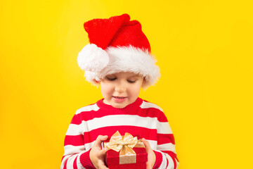Funny child toddler in a Santa hat with red gift , yellow background. Holidays, Traditions, Christmas concept.
