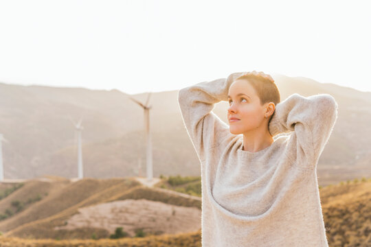 Shaved woman with hand behind head standing at wind farm