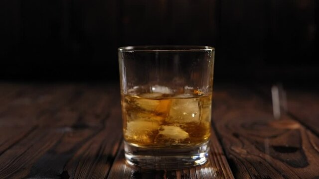 Close-up of ice cubes falling into a glass of whiskey or cognac on a wooden background, a bright light illuminates the glass creating Golden highlights. Slow motion.