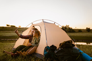 White couple taking selfie on cellphone and sitting in tent during camping