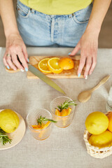 Female hand cut orange, Woman preparing, making citrus and rosemary fresh lemonade in glass on a white table at home, summer drink, detox healthy water.