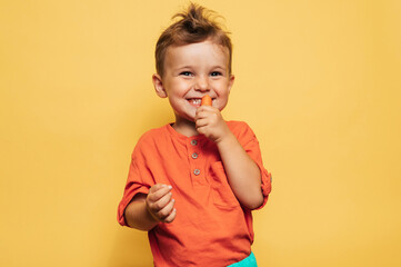 Studio shot of a smiling boy eating fresh carrots on a yellow background. The concept of healthy...