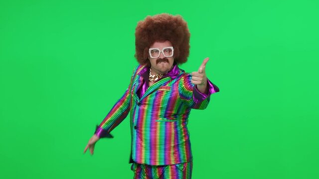 Retro dude in a funny wig and multicolored suit is dancing a funny spinning dance on a green background, a festive mood, a retro disco man is having fun at a new year's party.