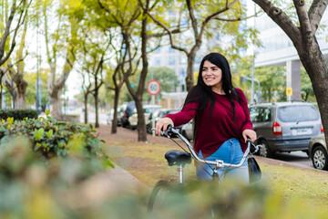 Fototapeta na wymiar young latina female student walking down the sidewalk with her bike in hand, while looking smiling at the route. copy space