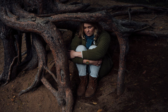 Lost woman sitting under tree roots while hugging knees