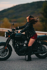 Fototapeta na wymiar Portrait of confident motorcyclist woman in mini skirt, leather jacket sits on retro-styled motorcycle. Attractive driver in jackboots on highway. Trip, speed, style, feminism concept.