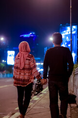 young couple holding hand while walk at road side of night city jakarta.